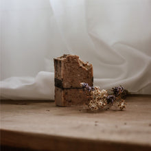 Load image into Gallery viewer, Savon exfoliant en pain, brun foncé, posé sur une surface avec fleurs séchées