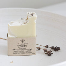 Load image into Gallery viewer, Savon en pain crème sur boîte ZARITS, avec fleurs et herbes séchées à côté.
